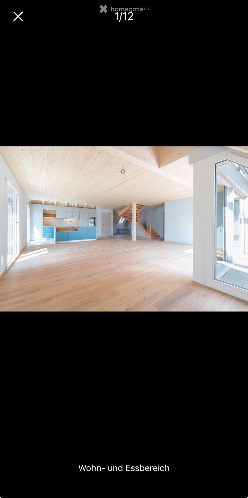 Open-plan living and dining area with light wooden flooring, exposed wooden ceiling beams, and modern kitchen cabinets. A wooden staircase leads to an upper level.