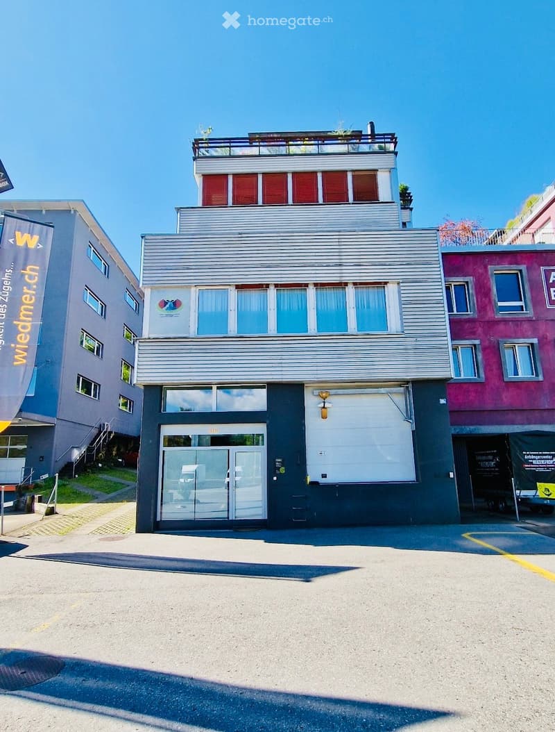 Modern three-story building with a combination of grey metal siding and red shutters, featuring a large glass entrance and a rooftop terrace with plants.