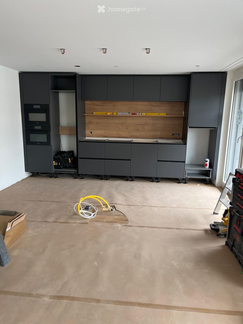 Empty room with grey cabinets, exposed wooden shelf, and construction tools scattered around. Bare concrete floor and white walls.