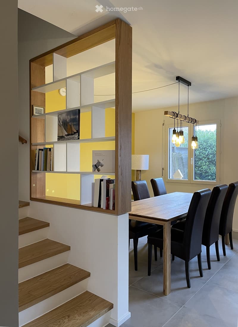 Furnished Room. Modern dining area with a wooden table, black chairs, a built-in bookshelf, and a hanging light fixture.