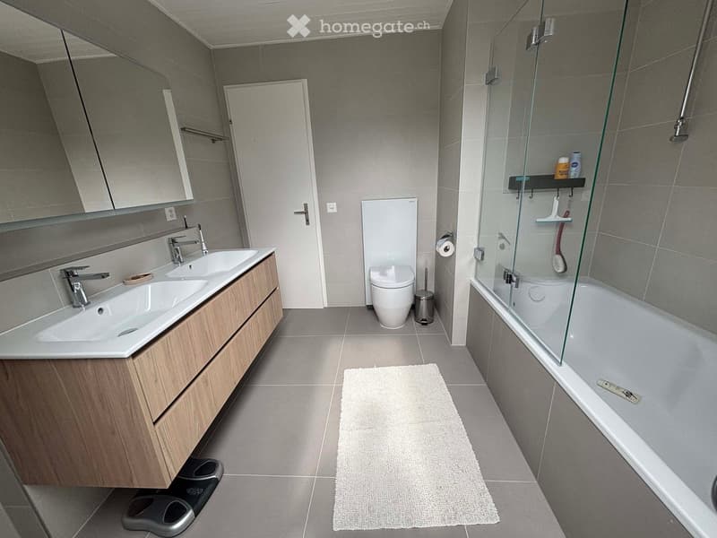 Bathroom. Modern bathroom with double sink, wooden vanity, bathtub with glass enclosure, toilet, and a small rug on the floor.