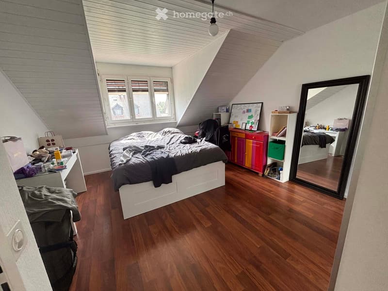 Bedroom. A cozy attic bedroom with wooden flooring, a large bed, a colorful cabinet, a large mirror, and a shelf with books and items.