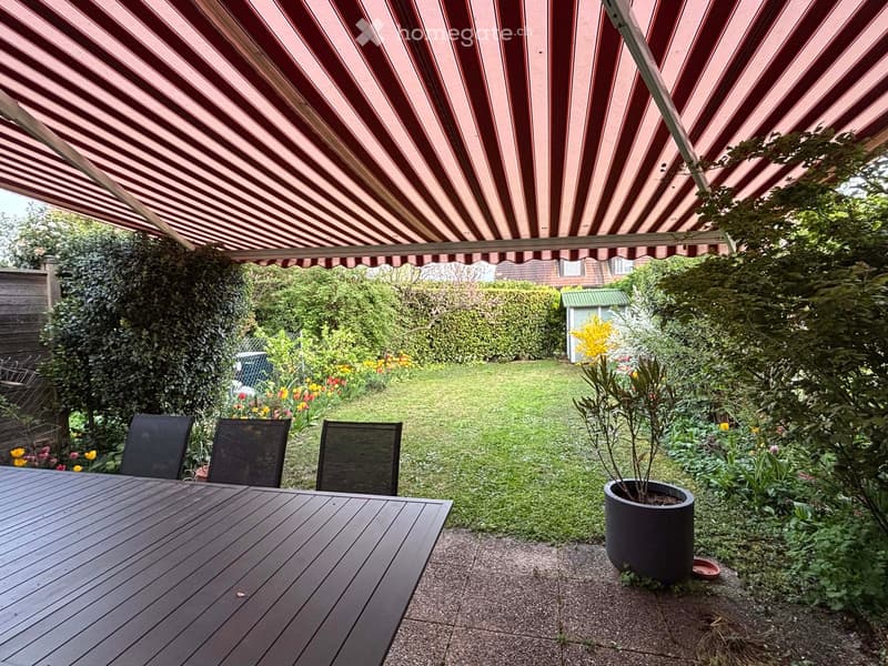 Garden / Outdoor Area. Garden area with a wooden table, three chairs, potted plant, and a striped awning providing shade.