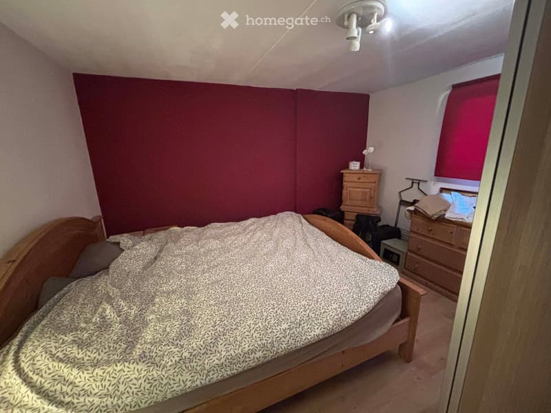 Bedroom. A cozy bedroom with a wooden bed, patterned bedspread, nightstand, and a small wooden drawer unit. The walls are painted in a deep maroon color, and there is a window with red blinds.