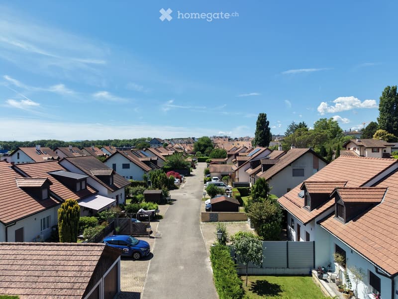 Residential neighborhood with multiple houses featuring red-tiled roofs, surrounded by greenery and parked cars.