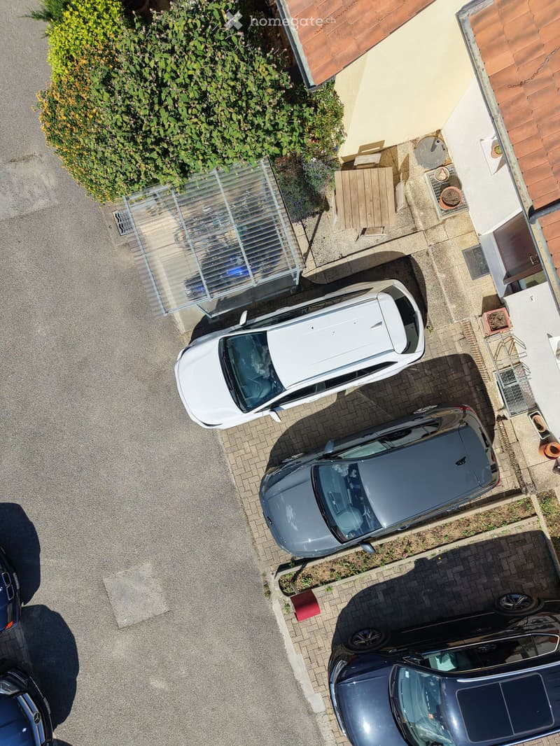 Aerial view of a residential driveway with three parked cars, a small wooden table, and a fenced garden area with greenery.