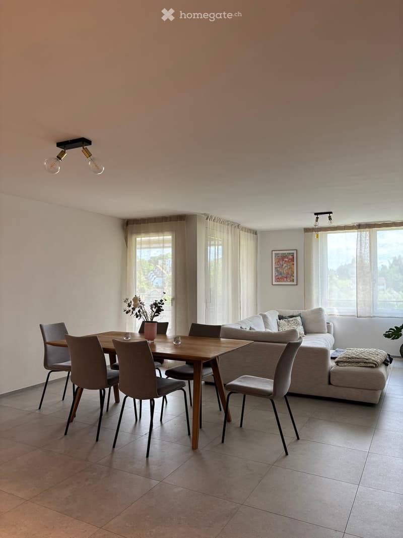Modern living and dining area with a sofa, dining table, chairs, and decorative vase. Large windows with sheer curtains and a view of greenery.