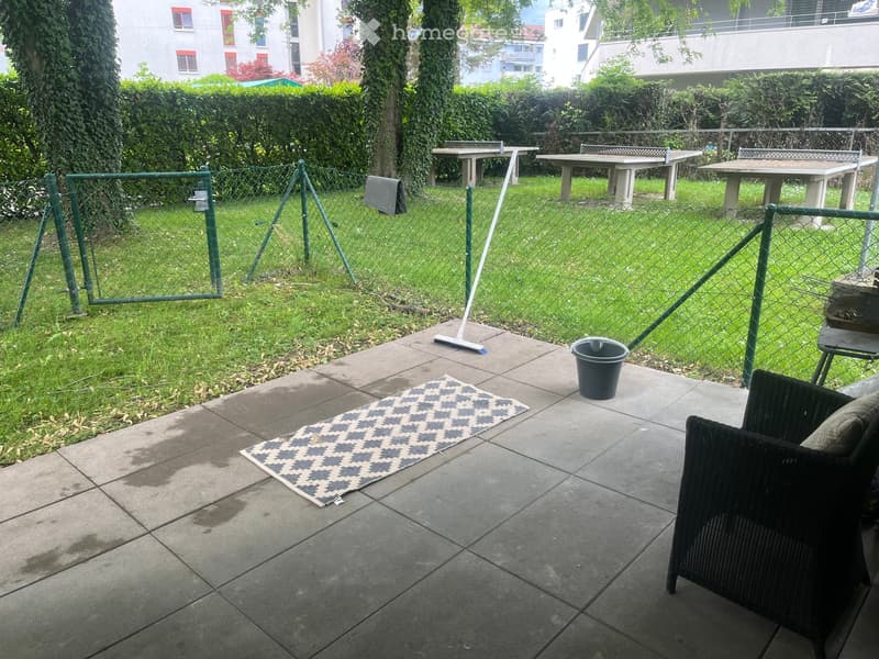 Outdoor patio area with tiled floor, a black wicker chair, a small rug, a broom, and a bucket. A fenced yard with tables and greenery is visible in the background.