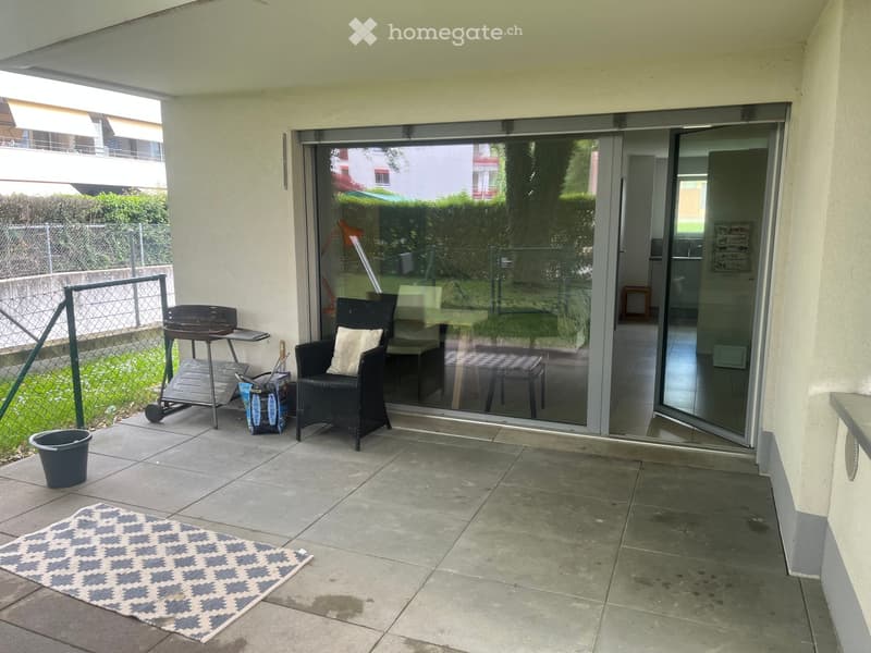 Balcony with tiled floor, a wicker chair with a cushion, a small table, a barbecue, and a patterned rug.