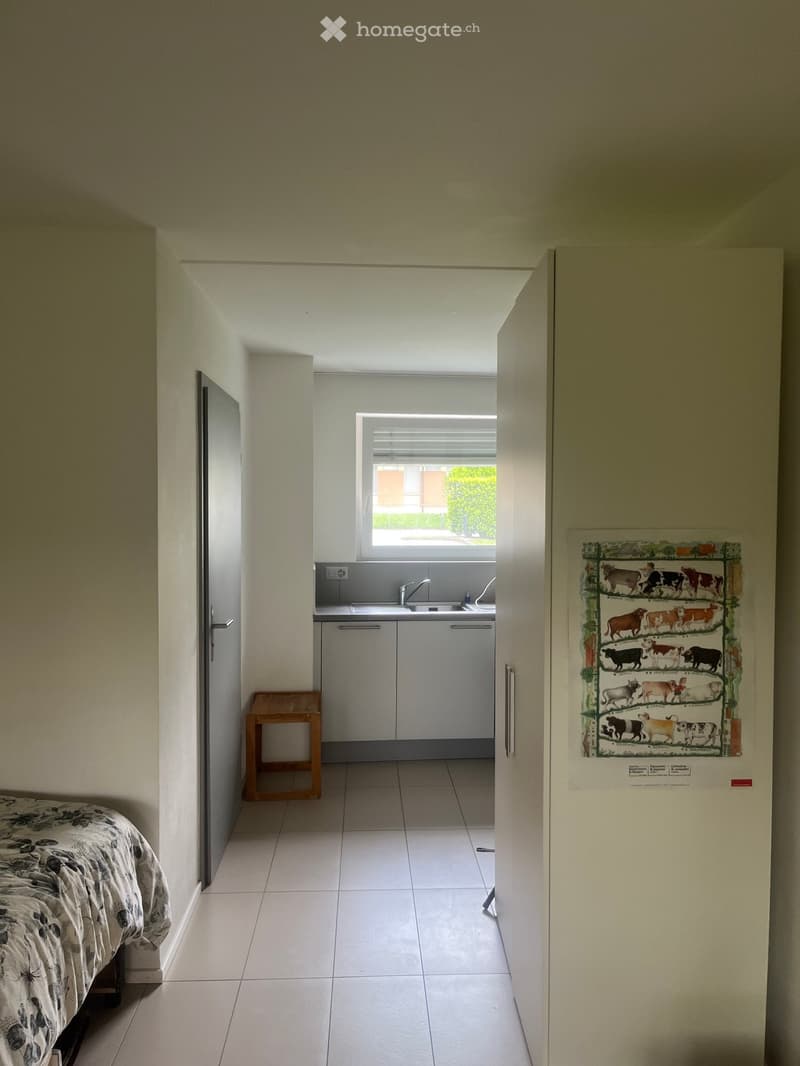 A furnished room with a bed, a small wooden stool, a refrigerator with a decorative poster, and a small kitchen area with a sink and faucet.