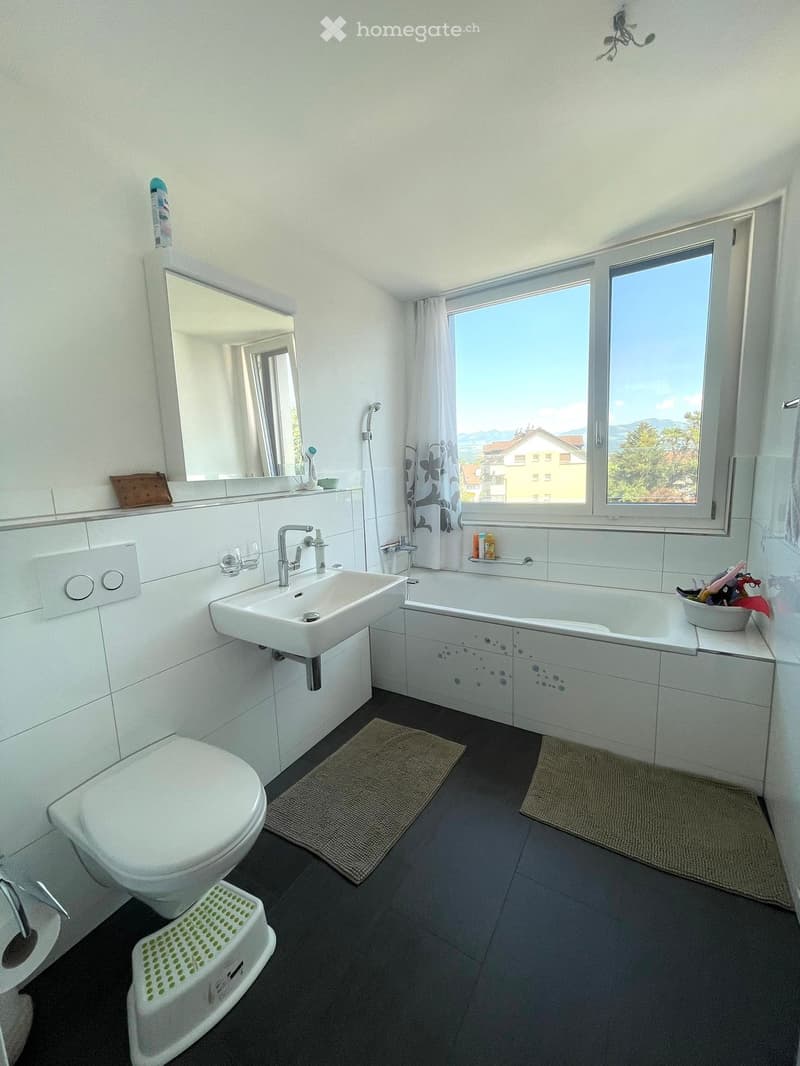 Modern bathroom with white tiles, a large window, a sink, a toilet, a bathtub, and a mirror. The floor is dark grey with two beige bathroom mats placed in front of the sink and bathtub.