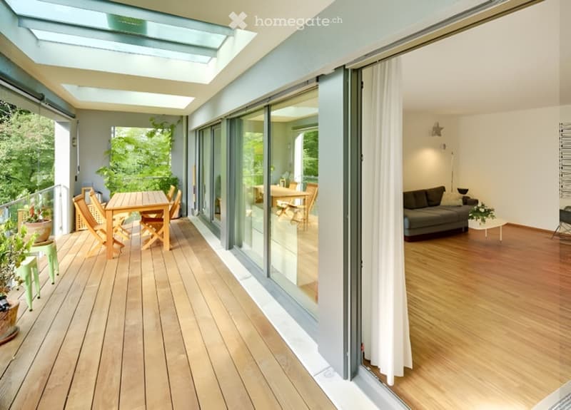 Wooden deck with a dining table and chairs, large glass sliding doors leading to a living room with a sofa and coffee table, and a view of greenery.