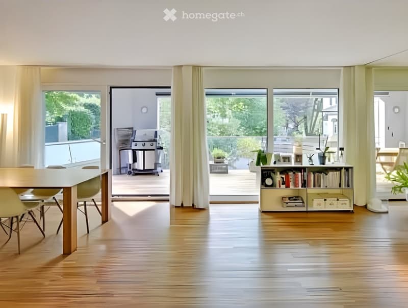 Bright, open-plan living and dining area with wooden flooring, large windows, and a balcony. Includes a dining table with chairs, a bookshelf, and potted plants.