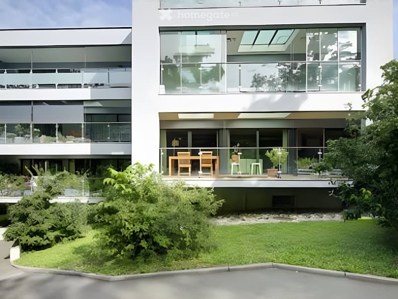 Modern multi-story building with large glass windows and balconies, surrounded by a well-maintained garden and lawn.