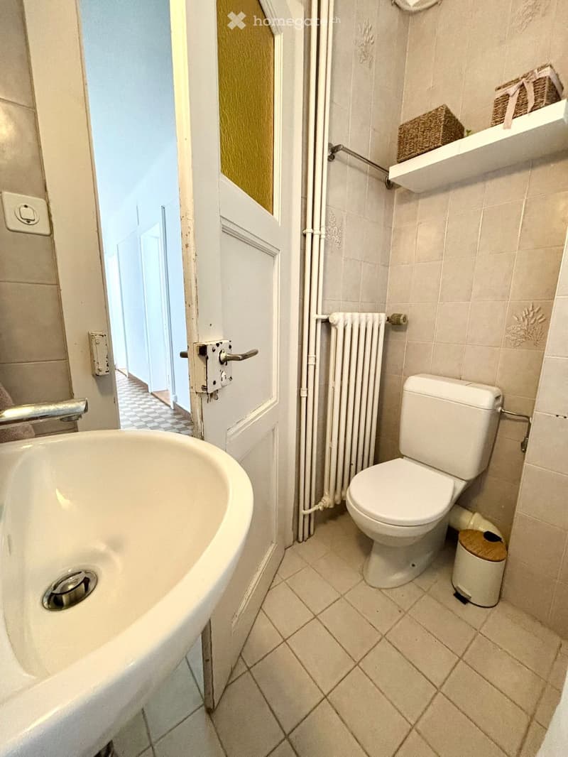 A small bathroom with a bathtub, toilet, radiator, and a trash bin. The walls are tiled, and there is a shelf with decorative items visible in the background.