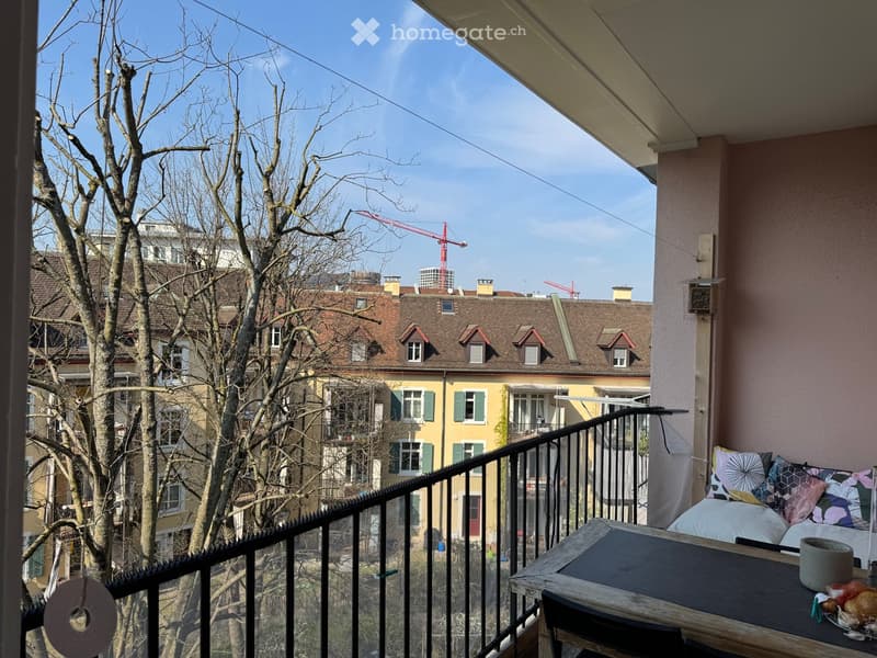 Balcony with a small table, chair, and decorative pillows, overlooking a cityscape with buildings and construction cranes.