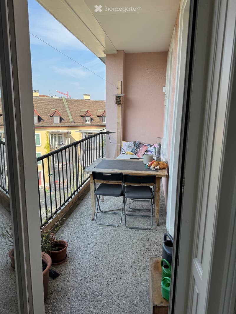 Balcony with a small table and two chairs, a cup, a bowl, and potted plants. The balcony has a view of neighboring rooftops.