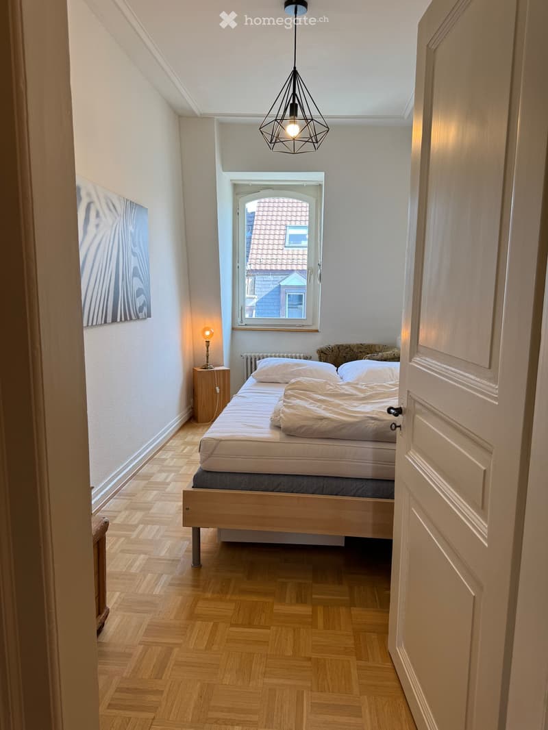 Cozy bedroom with wooden flooring, a single bed with white bedding, a nightstand with a lamp, and a picture frame on the wall. A window offers a view of a neighboring building.