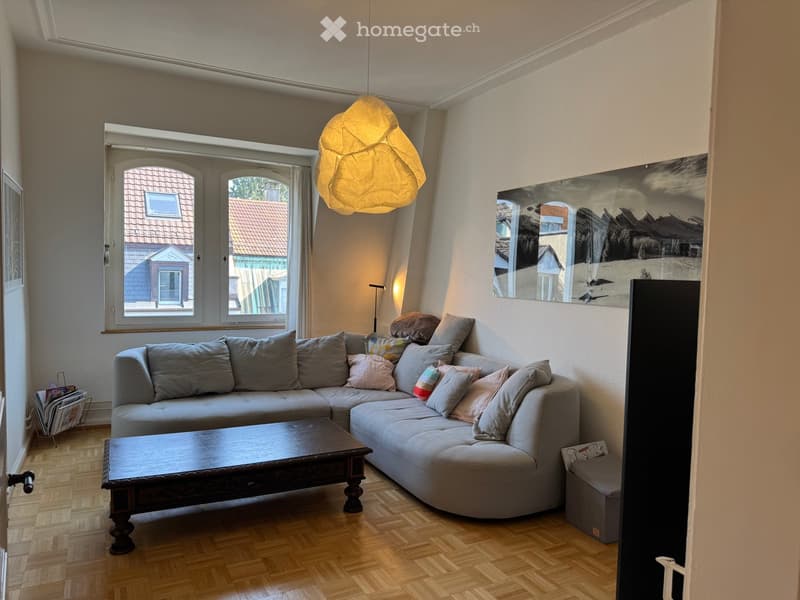 Living room with a grey sectional sofa, wooden coffee table, floor lamp, large mirror, and artwork on the wall. The room features wooden flooring and large windows with a view of the street.