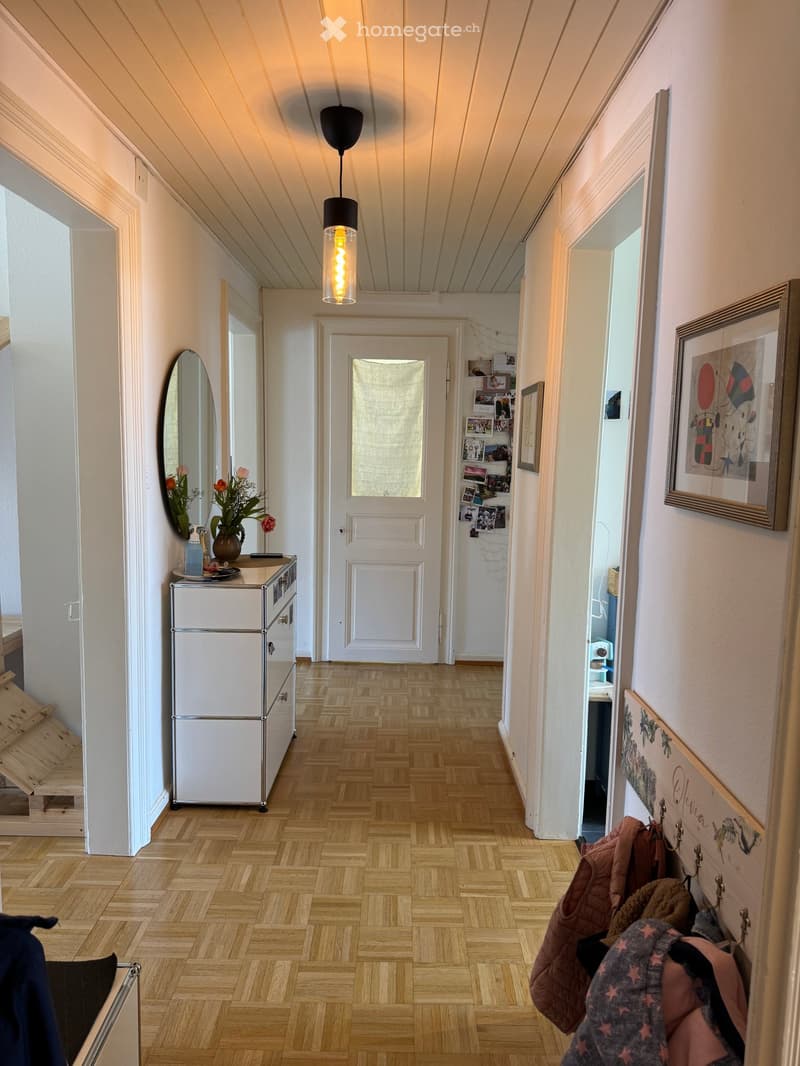 A well-lit hallway with wooden flooring, white walls, and a hanging pendant light. Features include a small cabinet with a vase of flowers, a mirror, framed artwork, and a coat rack with jackets.