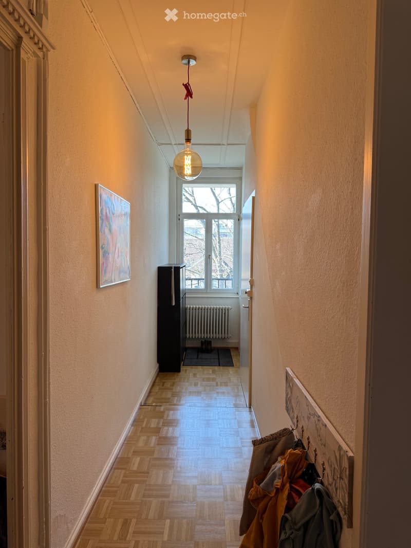 Hallway with wooden herringbone flooring, warm lighting, artwork on walls, coat rack with jackets, and a window at the end.