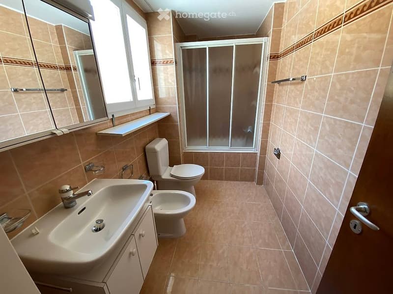 Bathroom with beige tiles, a sink with a faucet, a toilet, a bidet, a shower with a glass door, and a mirror above the sink.