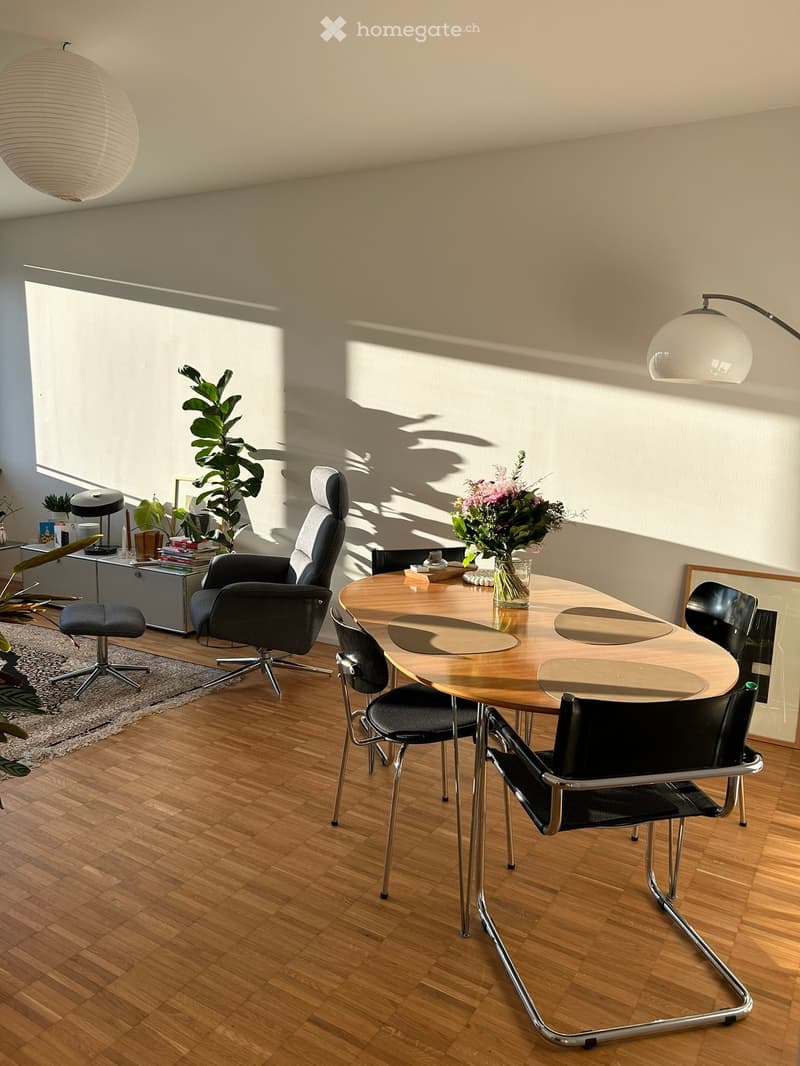 Modern dining area with a round wooden table, black chairs, a grey leather chair, a floor lamp, a plant, and decorative items on a side table.