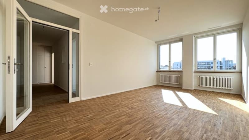Empty room with wooden flooring, large windows, and a glass door leading to another space.