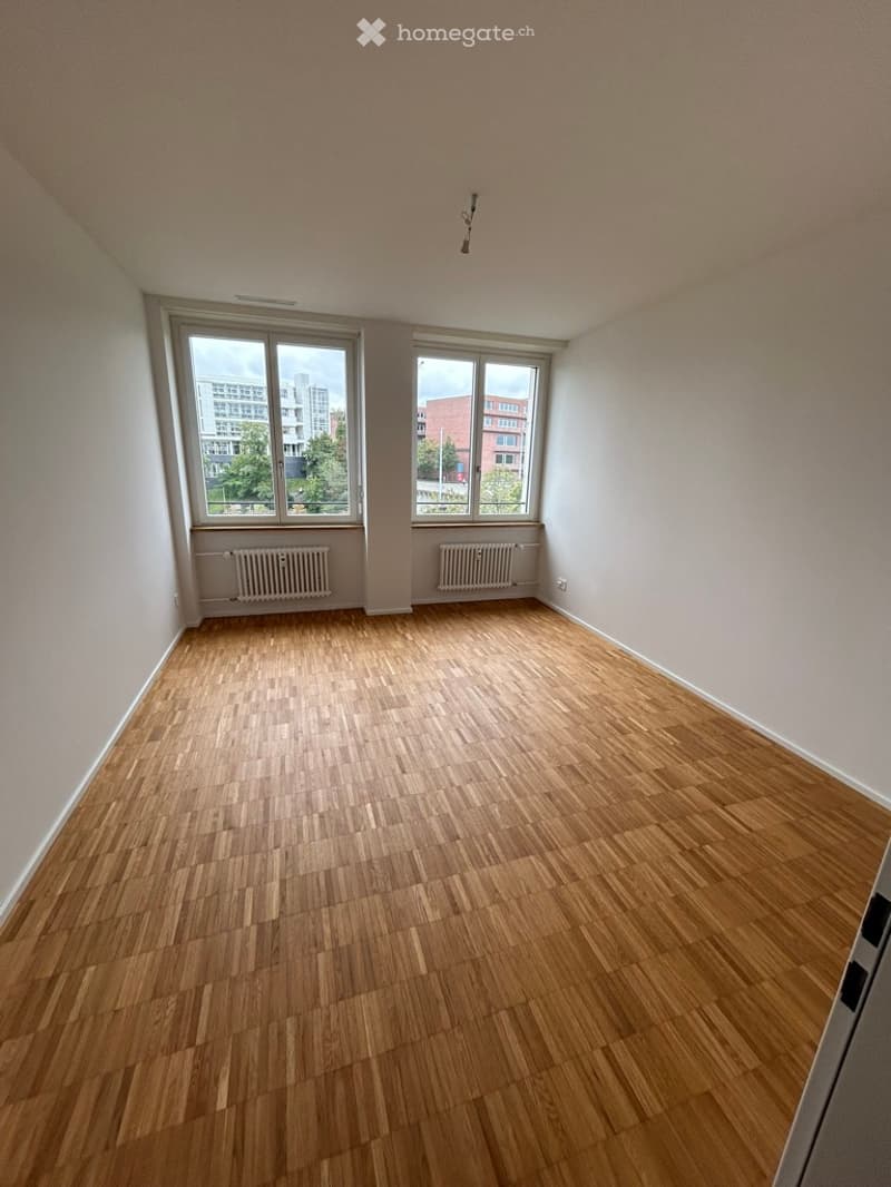 Empty room with wooden flooring, white walls, and large windows offering a view of buildings and greenery outside.