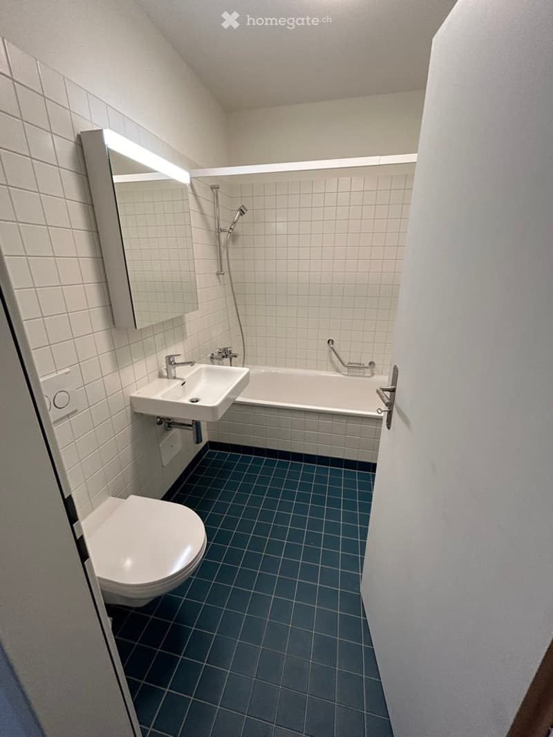 Modern bathroom with white tiles, blue floor tiles, a toilet, sink, bathtub, and a mirror with built-in lighting.