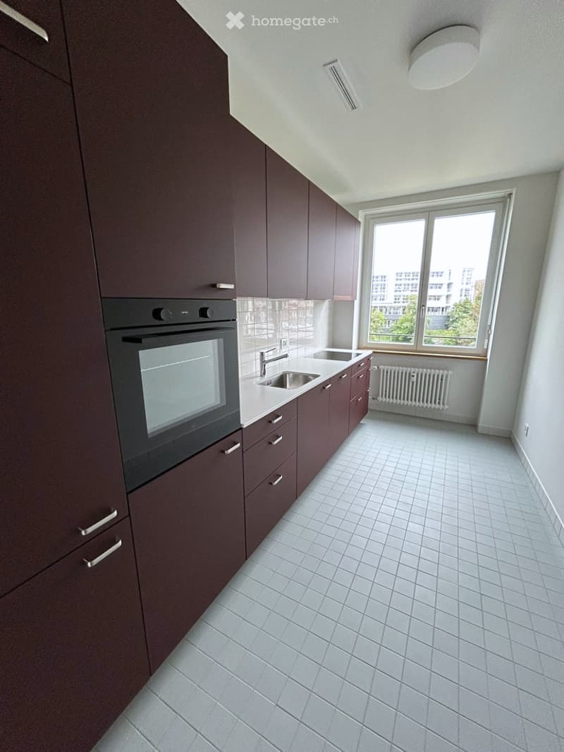 Modern kitchen with dark brown cabinets, white tiled floor, built-in oven, sink, and faucet. Large window provides natural light.