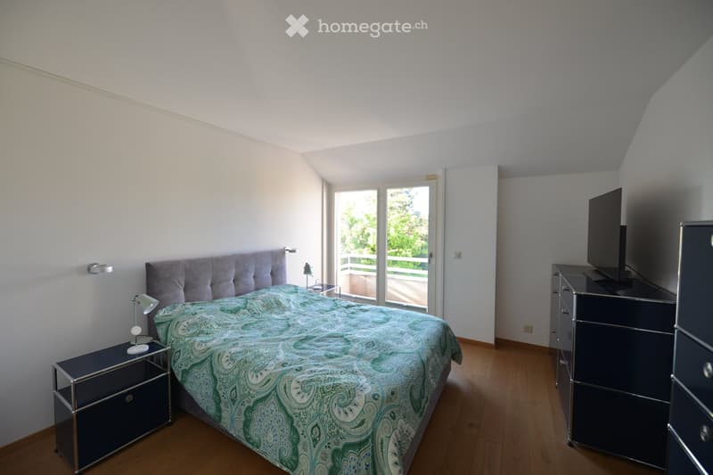 Spacious bedroom with wooden floor, white walls, and a large bed with a paisley bedspread. A nightstand with a lamp is on the left, and a dresser with a TV is on the right. A sliding glass door leads to a balcony.