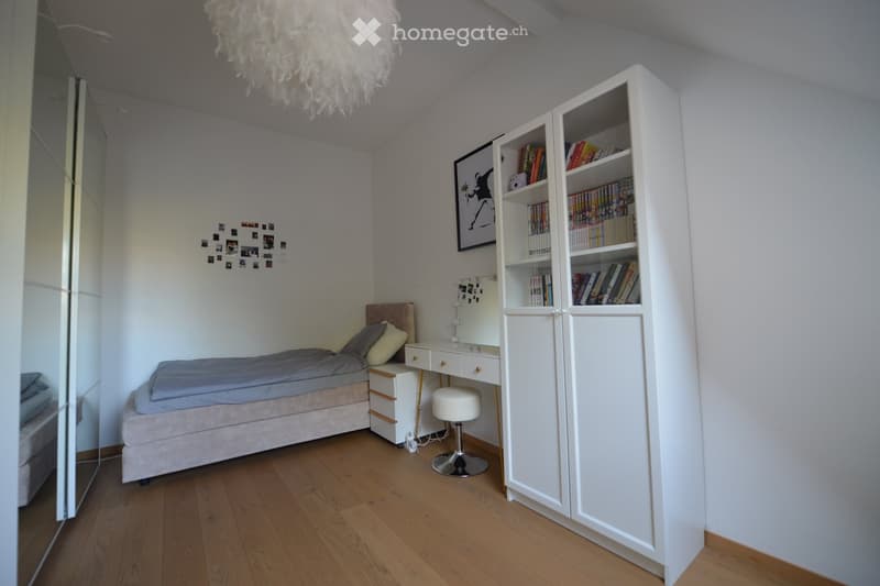A bedroom with a bed, desk, stool, bookcase, and a mirror. The room has a white ceiling, wooden floor, and white walls. The bed has a blue blanket and pillows. The desk has a drawer and a stool. The bookcase has books and a picture frame. The room has a l
