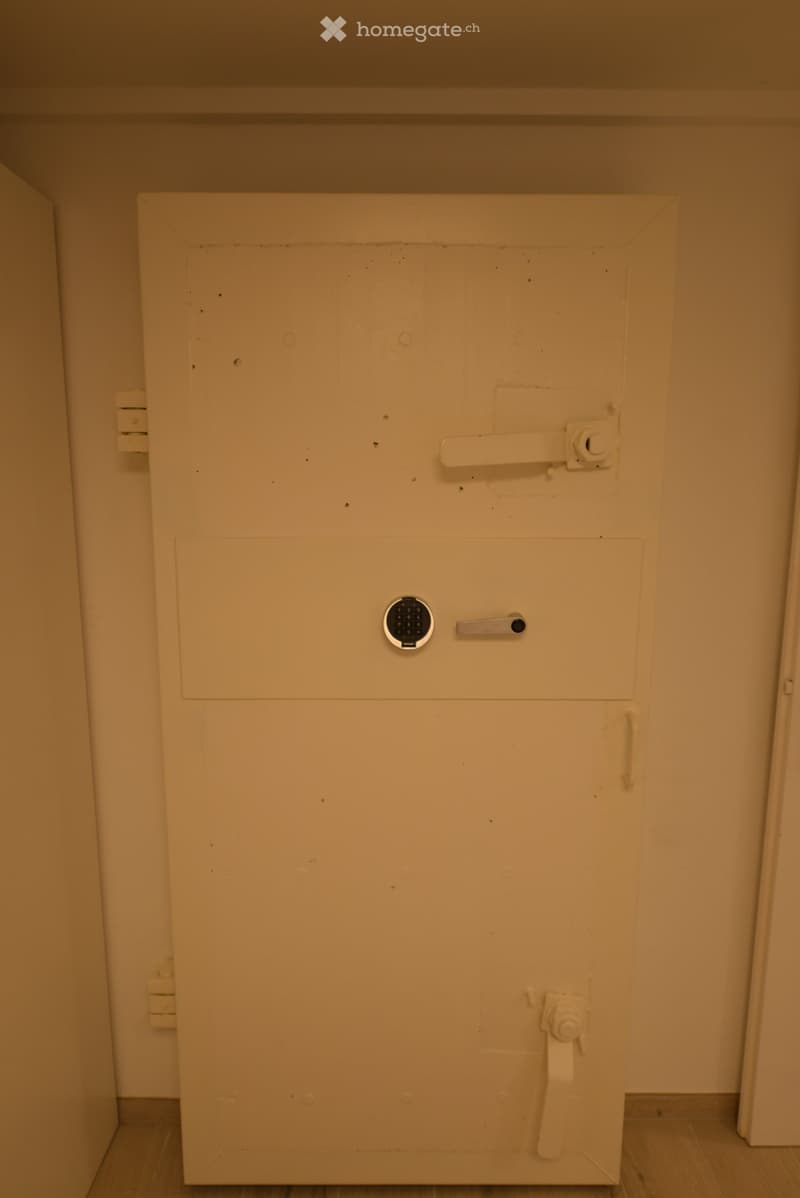 An empty room with a large white door, a keypad, and a handle. The door is closed, and there is a small hole in the middle of the door.