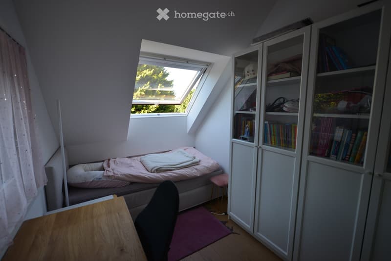 Small bedroom with a bed, desk, chair, and a wardrobe. The bed has a blanket and a pillow. The desk has a chair. The room has a window and a rug. The wardrobe is filled with books and a stuffed toy.