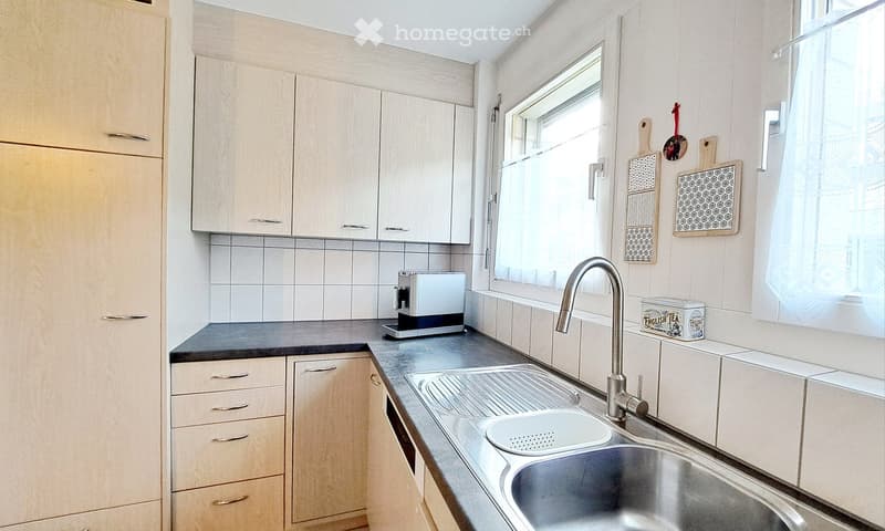Kitchen. Modern kitchen with light wooden cabinets, stainless steel sink, and a toaster on the counter. The window has a white curtain, and decorative cutting boards are mounted on the wall.