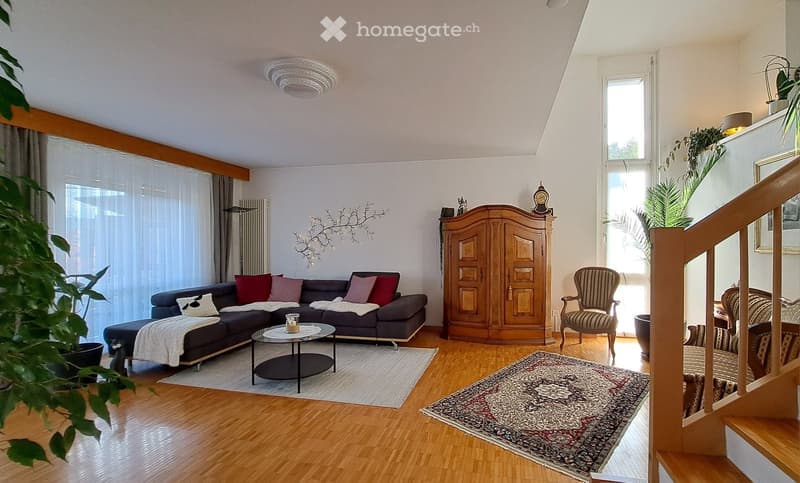 Living Room. Living room with wooden flooring, large windows, a dark grey sectional sofa with colorful cushions, a round black coffee table, a wooden cabinet, a patterned rug, and a decorative plant in the corner.