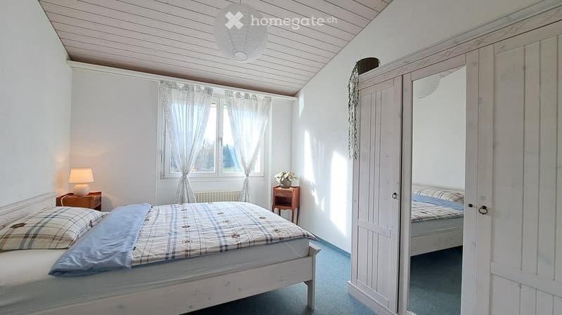 Bedroom. Bright bedroom with a wooden bed, plaid bedding, nightstand with lamp, open wardrobe with mirror, and a small wooden side table with a flower vase. Natural light filters through sheer curtains.