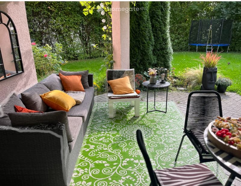 Outdoor patio with green patterned rug, grey sofa with orange cushions, white chair, small round table, black wicker chair, and decorative items.