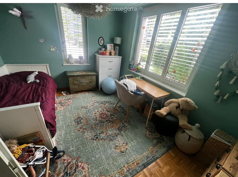 Cozy bedroom with a bed, a chair, a small table, a stool, and a cat. The room features large windows with blinds, a patterned rug, and various decorative items including a dreamcatcher and a small table with a lamp.