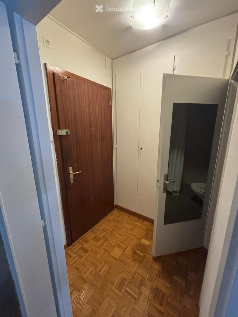 A narrow hallway with wooden flooring, white walls, and a wooden door on the left leading to another room. A partially open door on the right reveals a bathroom with a toilet and a mirror.