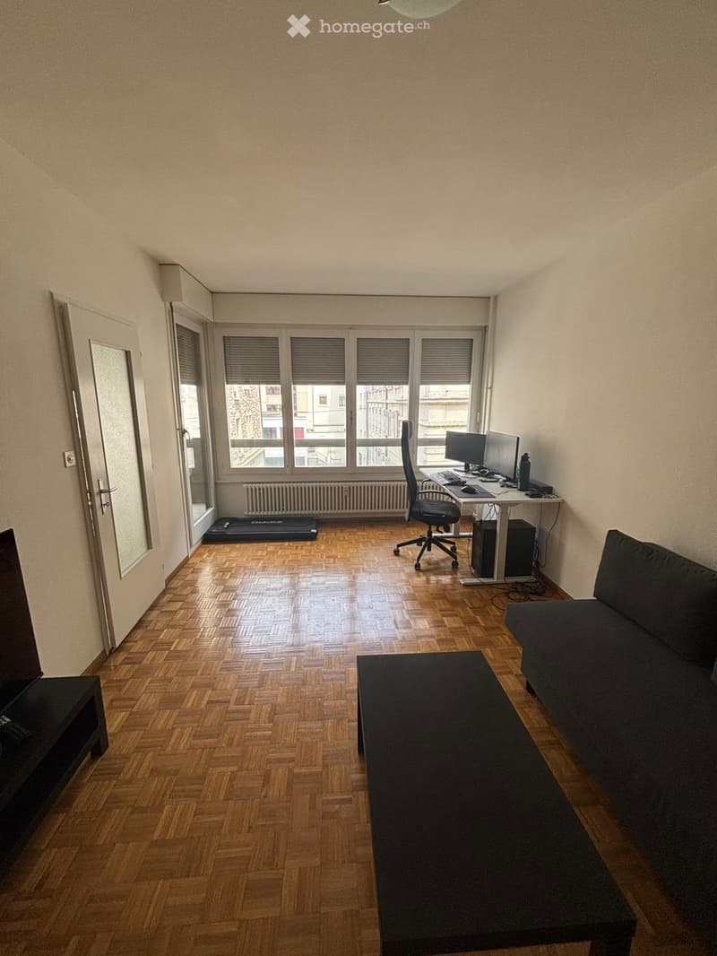 A modern, multi-functional room with wooden flooring, large windows, a sofa, coffee table, desk with computer, and exercise mat.