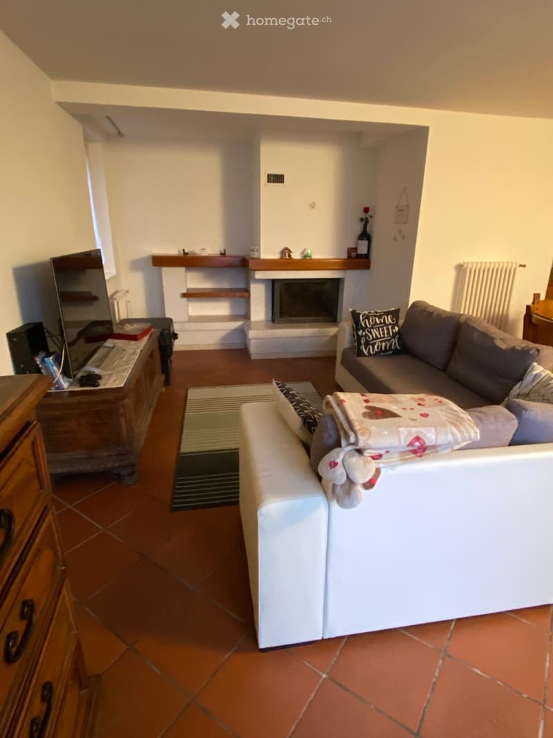 Living room with a fireplace, a grey sofa, a white ottoman, a wooden cabinet, and a television. The room features a tiled floor and a rug in front of the sofa.