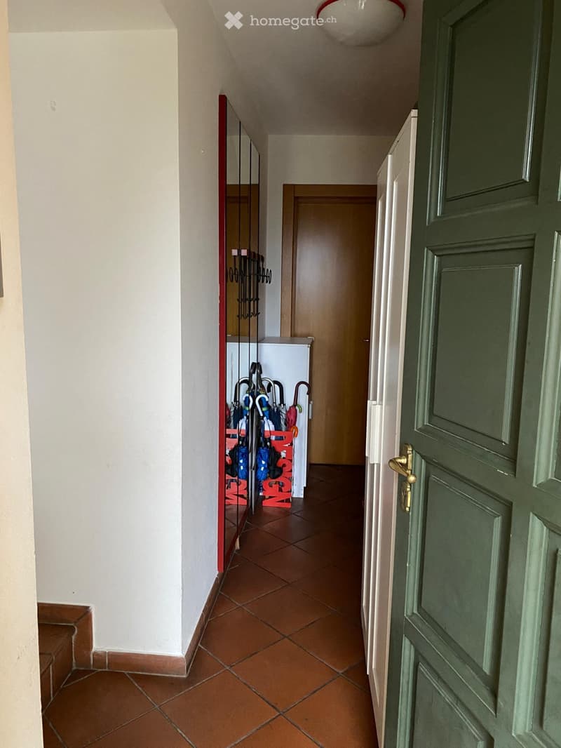 A narrow hallway with tiled flooring, white walls, and a coat rack holding umbrellas. A green door is visible on the right side.