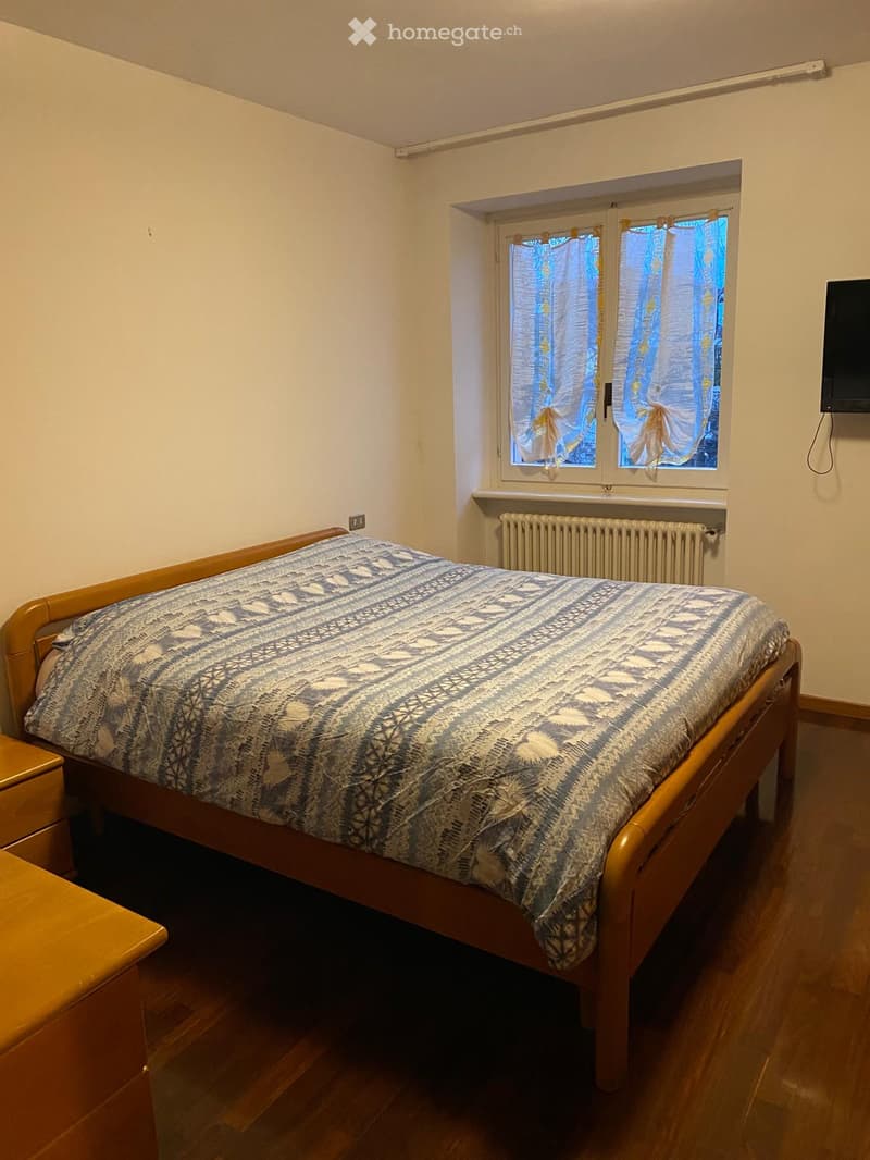 Cozy bedroom with wooden bed, patterned bedspread, nightstand, radiator, and TV mounted on wall. Light-colored walls and wooden flooring.