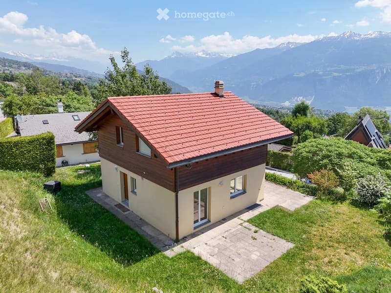 Single-story house with red-tiled roof, wooden siding, and large windows, situated in a green landscape with mountain views.