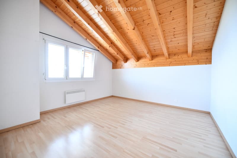 Empty attic room with wooden slanted ceiling, light wooden flooring, white walls, and a window with white frame. A white radiator is mounted on one wall.