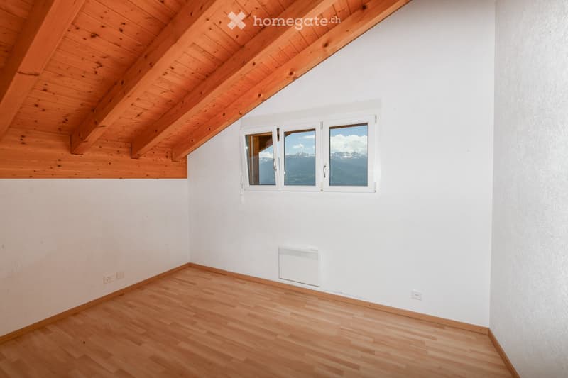 Empty room with wooden floor, white walls, wooden slanted ceiling, and large window offering a mountain view.
