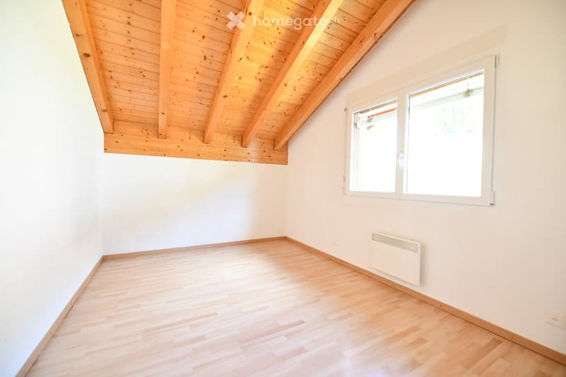 Empty room with light wooden flooring, white walls, and a slanted wooden ceiling. Two windows on one wall and a radiator below them.