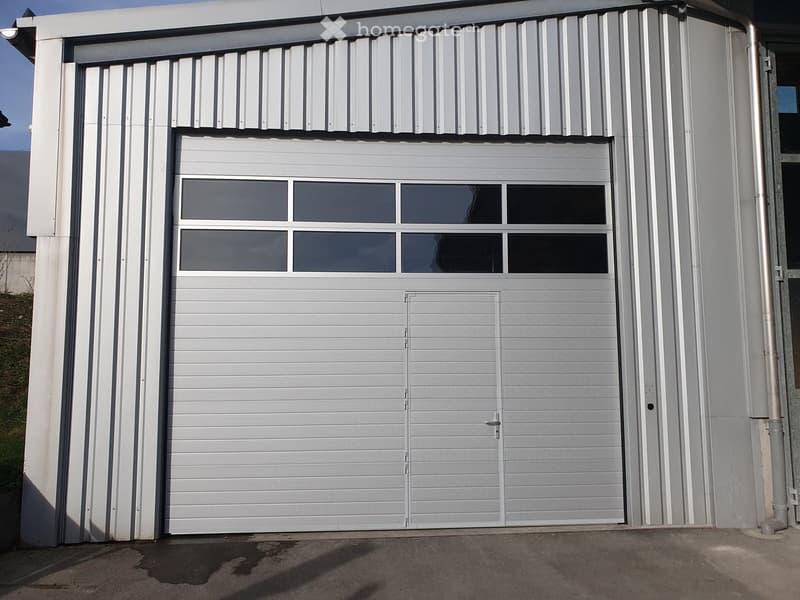 A modern, grey garage with a large, closed garage door featuring a window section at the top. The garage has a sleek, contemporary design with clean lines and a metallic finish.